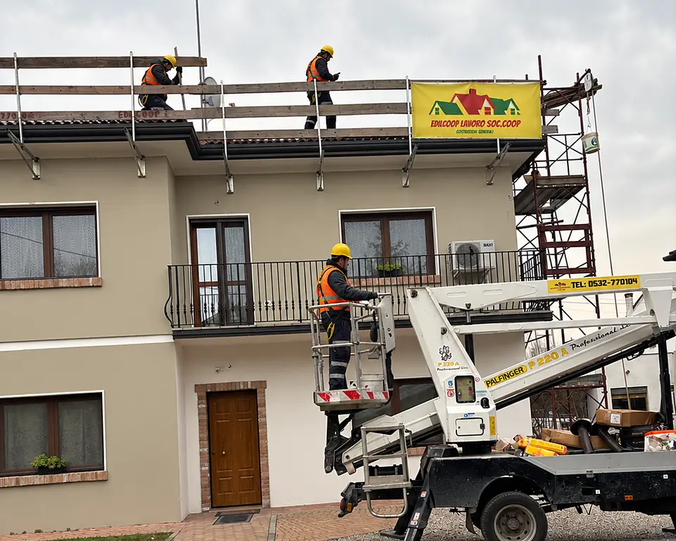 Operai Edilcoop con casco sul tetto protetto da parapetti e un operaio sulla piattaforma aerea, telone Edilcoop sulla facciata